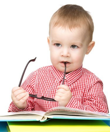 Cute little child play with book and biting glasses while sitting at table, isolated over whiteの写真素材