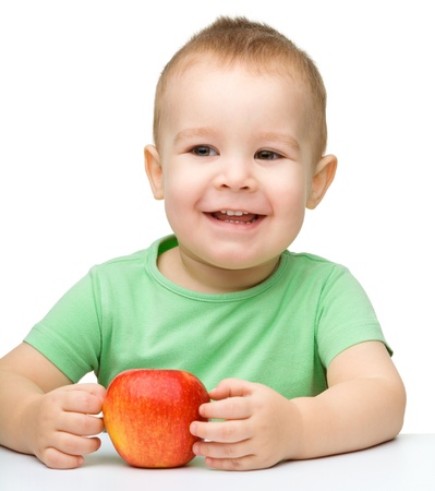 Portrait of a cute cheerful little boy, who is smiling while sitting at table with apple, isolated over whiteの写真素材