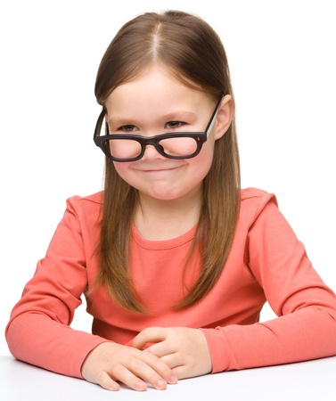 Happy little girl sits at a table and smile while wearing glasses, isolated over whiteの写真素材