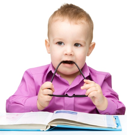 Cute little child play with book and biting glasses while sitting at table, isolated over whiteの写真素材