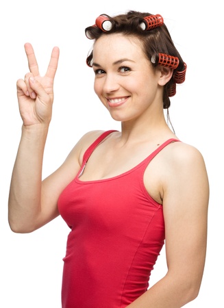 Young woman dressed in red is showing victory sign while wearing hair-rollers, isolated over whiteの写真素材