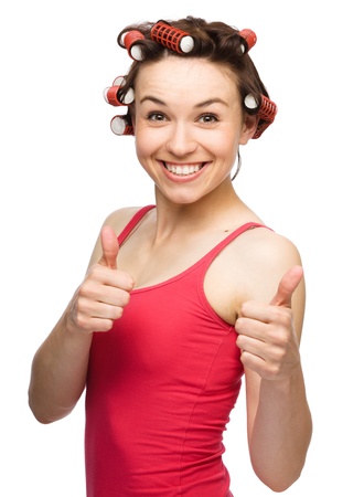 Young woman dressed in red is showing thumb up gesture using both hands while wearing hair-rollers, isolated over whiteの写真素材