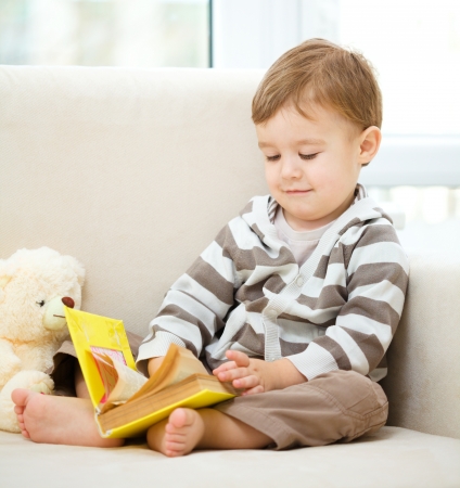 Cute little boy is reading book while sitting on a sofaの写真素材