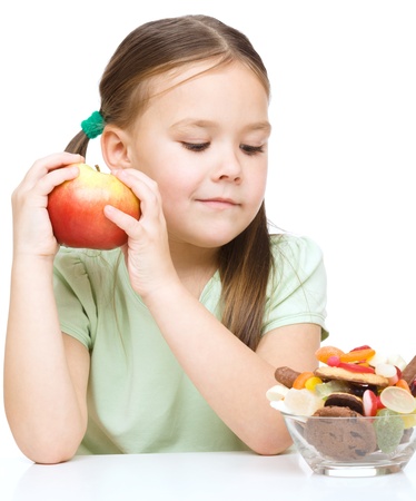 Cute little girl choosing between apples and sweets, isolated over whiteの写真素材