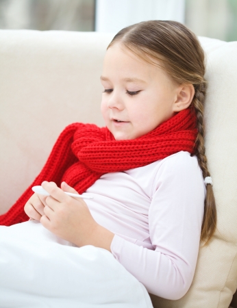Ill little girl is reading thermometer while laying on sofaの写真素材