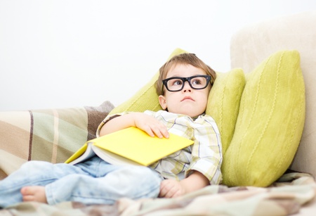 Cute little child holding a book and wearing glasses while laying on couchの写真素材