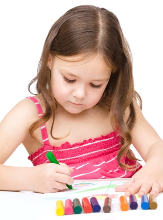 Little girl is drawing using a crayon while sitting at table, isolated over whiteの写真素材