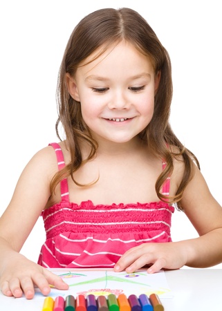 Little girl is drawing using colorful crayons while sitting at table, isolated over whiteの写真素材