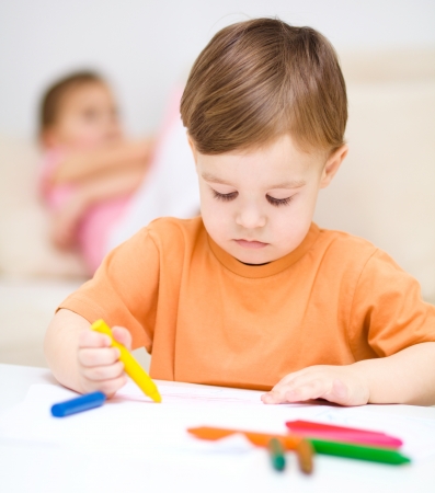 Little boy is drawing on white paper using crayon, his sister sits behind himの写真素材