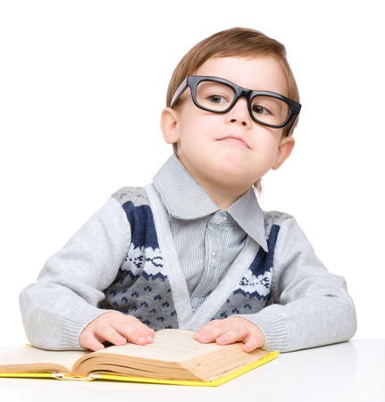 Cute little child play with book and wearing glasses while sitting at table, isolated over whiteの写真素材