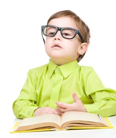 Cute little child play with book and wearing glasses while sitting at table, isolated over whiteの写真素材