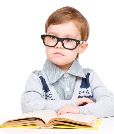 Cute little child play with book and wearing glasses while sitting at table, isolated over whiteの写真素材