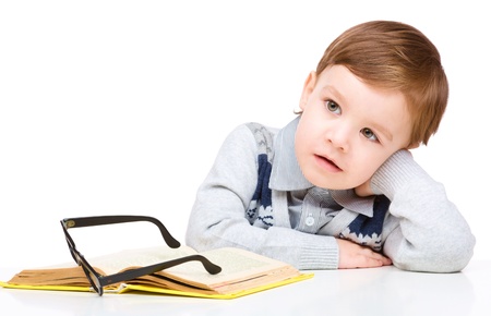 Cute little child play with book and wearing glasses while sitting at table, isolated over whiteの写真素材