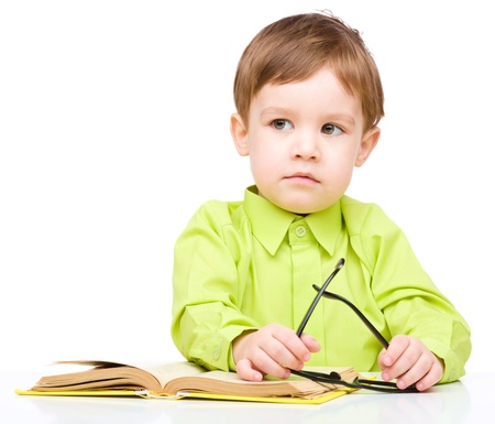 Cute little child play with book and wearing glasses while sitting at table, isolated over whiteの写真素材
