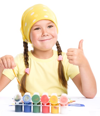 Cute cheerful child play with paints while sitting at table and showing thumb up gesture, isolated over whiteの写真素材