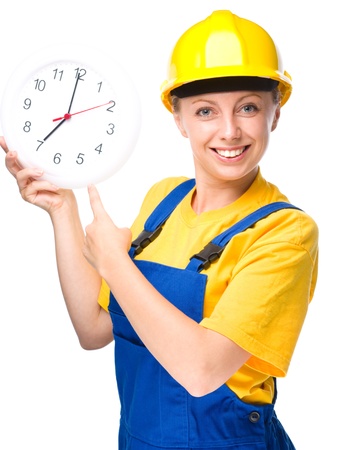 Young happy lady as a construction worker is pointing at clock showing seven, get up early concept, isolated over whiteの写真素材