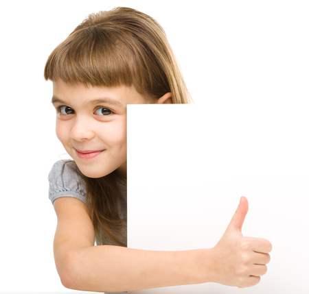 Little girl is looking out from the blank banner and showing thumb up sign, isolated over whiteの写真素材