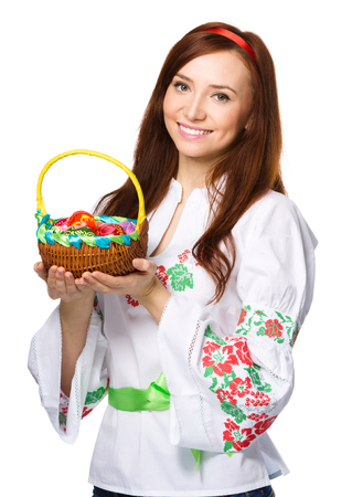 Young woman in ukrainian national cloth is holding a basket full of easter eggs, isolated over whiteの写真素材