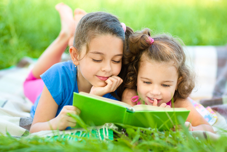 Two little girls are reading book while laying on green grass, outdoor shootの写真素材