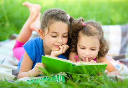 Two little girls are reading book while laying on green grass, outdoor shootの写真素材