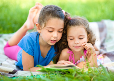 Two little girls are reading book while laying on green grass, outdoor shootの写真素材