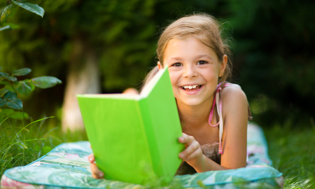 Cute little girl is reading a book while laying on green grassの写真素材