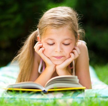 Cute little girl is reading a book while laying on green grassの写真素材