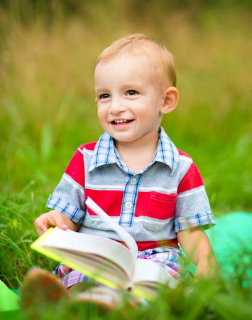 Little boy is reading book while sitting on green grass outdoorsの写真素材