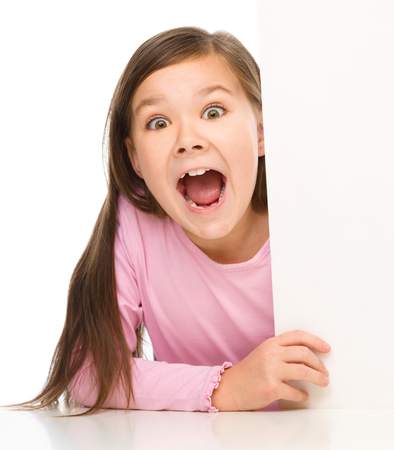 Little girl is looking from out blank board and showing thumb up sign, isolated over whiteの写真素材