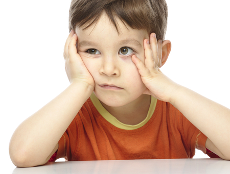 Portrait of a cute little boy sitting at table supporting his head with hands, isolated over whiteの写真素材