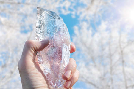 Piece of ice in a man's hand against the background of Winter trees and the bright sun. Global warming concept. Seasonal weather.の写真素材