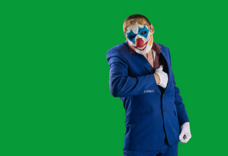 Man in a clown mask in a business suit takes out something dangerous from the inside pocket of his jacket. Isolated on a green background. Hand gesture in white gloves. Threat concept. Halloween.の写真素材