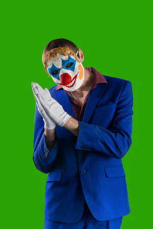 Man in a clown mask and a business suit. Isolated on a green background. The gesture of rubbing your hands. Halloween.の写真素材