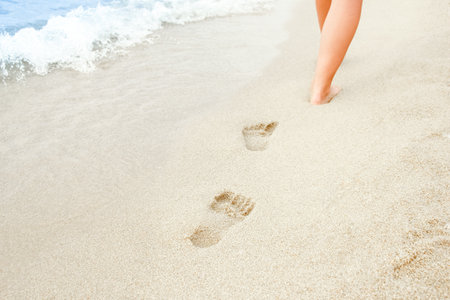Beach travel - woman relaxing walking on a sandy beach leaving footprints in the sand. Close up detail of female feet on golden sand at a beach in Greece. Background.の写真素材