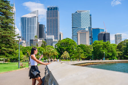 A young woman in a T-shirt and skirt stands in the park on the promenade and talks on the phone, skyscrapers in the business district, view from the Royal Botanic Garden Sydney, summer sunny day.の写真素材