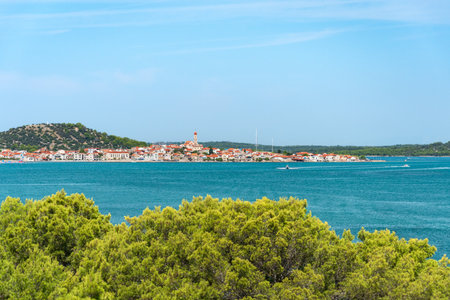 The coastal town of Tisno in Croatia is situated on the seashore, surrounded by low mountains and blue water. A view from the path of the summer landscape on a sunny day.の写真素材