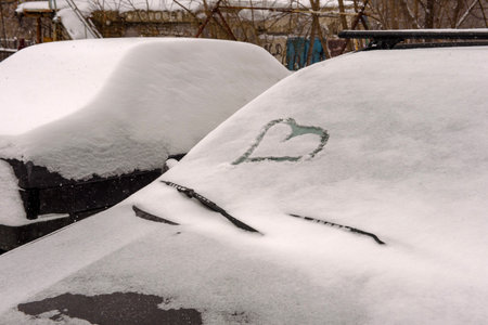A car covered in a layer of snow with a heart on the windshield, parked next to another snow-covered car.の写真素材