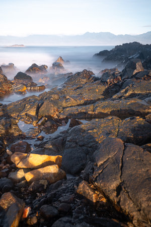 A breathtaking view of Diguisit Rock Formation at sunset, where rugged, weathered rocks in the foreground glow with warm orange hues. A small, tranquil pool reflects the serene beauty of the scene, while dramatic rock formations, including a pointed peak, rise majestically against the vibrant, colorful sky. The rocky coastline and panoramic horizon evoke a sense of tranquility and awe-inspiring natural wonder.の写真素材