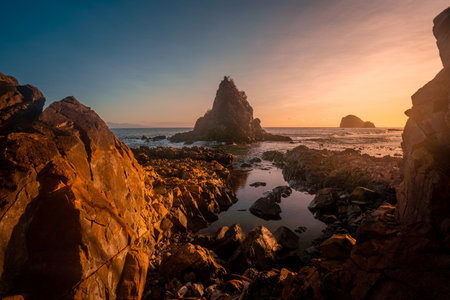 A breathtaking view of Diguisit Rock Formation at sunset, where rugged, weathered rocks in the foreground glow with warm orange hues. A small, tranquil pool reflects the serene beauty of the scene, while dramatic rock formations, including a pointed peak, rise majestically against the vibrant, colorful sky. The rocky coastline and panoramic horizon evoke a sense of tranquility and awe-inspiring natural wonder.の写真素材