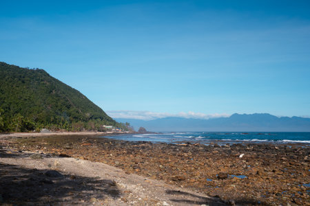 A scenic view of the coastline at Diguisit, Quirino, Philippines, featuring a rocky beach leading to the sea with a prominent island in the distance. Lush green vegetation covers the hillside on the right, contrasting with the rocky foreground. This image captures the natural beauty and tranquil atmosphere of this coastal location.の写真素材