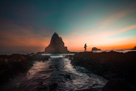 A breathtaking view of Diguisit Rock Formation at sunset, where rugged, weathered rocks in the foreground glow with warm orange hues. A small, tranquil pool reflects the serene beauty of the scene, while dramatic rock formations, including a pointed peak, rise majestically against the vibrant, colorful sky. The rocky coastline and panoramic horizon evoke a sense of tranquility and awe-inspiring natural wonder.の写真素材