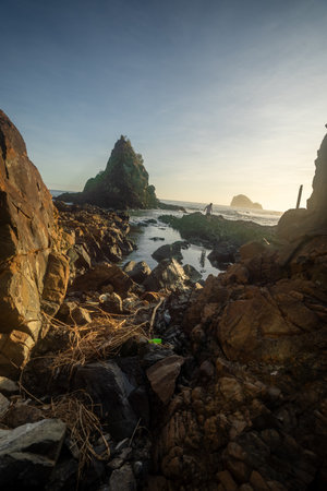 A breathtaking view of Diguisit Rock Formation at sunset, where rugged, weathered rocks in the foreground glow with warm orange hues. A small, tranquil pool reflects the serene beauty of the scene, while dramatic rock formations, including a pointed peak, rise majestically against the vibrant, colorful sky. The rocky coastline and panoramic horizon evoke a sense of tranquility and awe-inspiring natural wonder.の写真素材