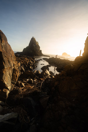 A breathtaking view of Diguisit Rock Formation at sunset, where rugged, weathered rocks in the foreground glow with warm orange hues. A small, tranquil pool reflects the serene beauty of the scene, while dramatic rock formations, including a pointed peak, rise majestically against the vibrant, colorful sky. The rocky coastline and panoramic horizon evoke a sense of tranquility and awe-inspiring natural wonder.の写真素材