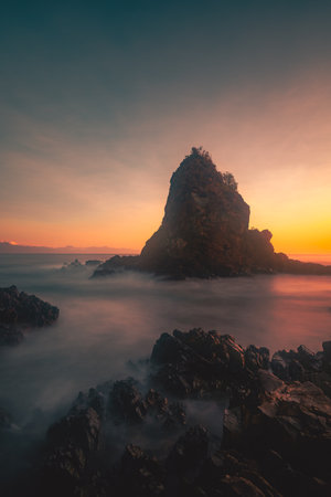 A breathtaking view of Diguisit Rock Formation at sunset, where rugged, weathered rocks in the foreground glow with warm orange hues. A small, tranquil pool reflects the serene beauty of the scene, while dramatic rock formations, including a pointed peak, rise majestically against the vibrant, colorful sky. The rocky coastline and panoramic horizon evoke a sense of tranquility and awe-inspiring natural wonder.の写真素材