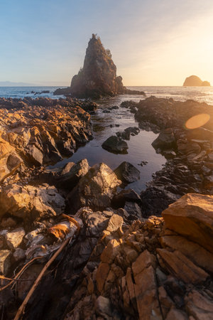A breathtaking view of Diguisit Rock Formation at sunset, where rugged, weathered rocks in the foreground glow with warm orange hues. A small, tranquil pool reflects the serene beauty of the scene, while dramatic rock formations, including a pointed peak, rise majestically against the vibrant, colorful sky. The rocky coastline and panoramic horizon evoke a sense of tranquility and awe-inspiring natural wonder.の写真素材