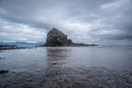 A dramatic coastal scene  of Diguisit, Aurora featuring two rugged rock formations on a sandy shore, under a sky filled with fast-moving, moody clouds. The calm water reflects the rich tones of the sky, creating a striking and atmospheric seascape.の写真素材