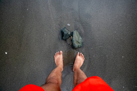 First-person perspective shot of bare feet standing on a dark, wet sandy beach. Two rocks are partially submerged in the shallow water near the feet. The bottom of bright red shorts is visible at the bottom of the frame. This image evokes feelings of relaxation, connection with nature, and simple pleasures.の写真素材