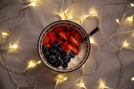 Top view of a bowl with yogurt, strawberries and blueberries over grey sheets with lightsの写真素材