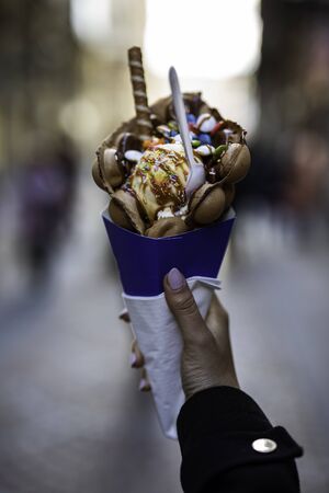 Woman hand holding a bubble waffle with ice cream and candies on a blue paper cone with blurred unrecognizable crowd on the background street in Europeの写真素材