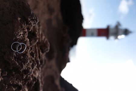 Focus on wedding rings on volcanic rock with blurred red and white lighthouse on the backgroundの写真素材